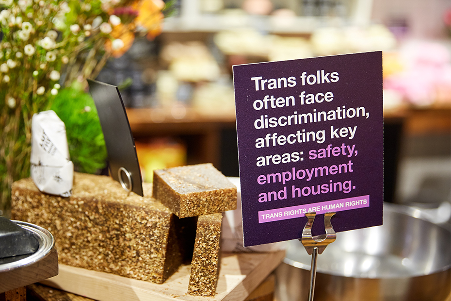 A purple point of sale sign stands beside a displace of naked products. The sign reads: Trans folks often face discrimination, affecting key areas: safety, employment and housing. Trans rights are human rights.