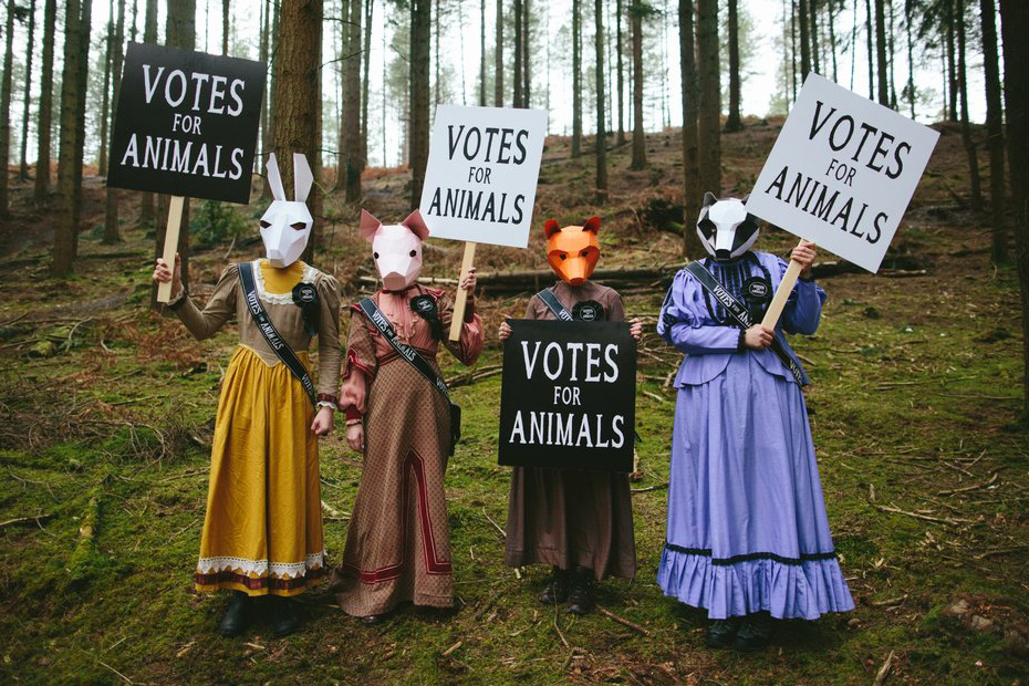 A group of four people stand in a field wearing animal masks, holding signs that read: votes for animals.