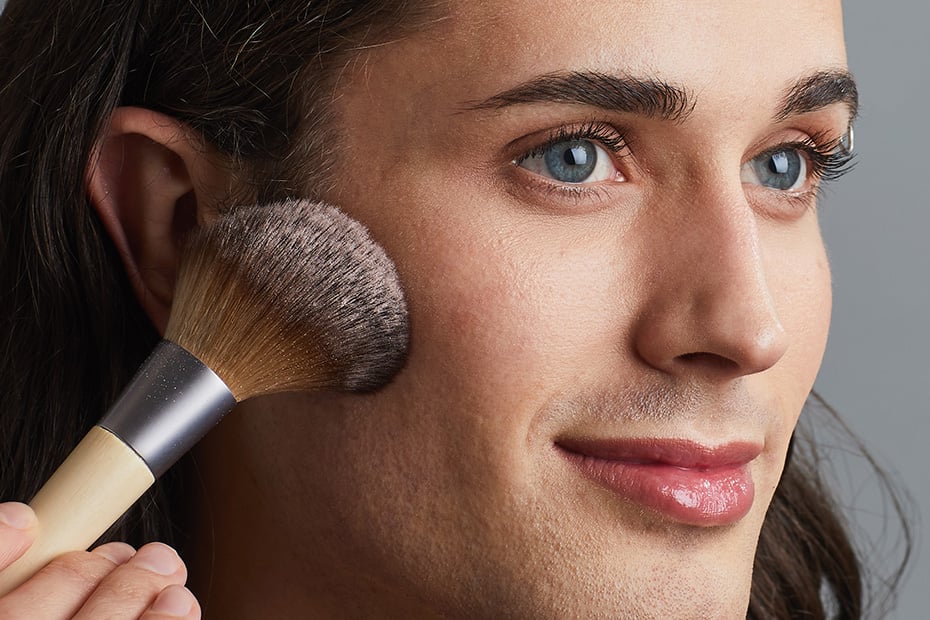 A close up shot of a model with brunette hair and blue eyes holding a gentle smile while looking off camera. They are holding a face powder makeup brush up to their cheek. The makeup brush has a light wood handle with aluminum detailing, and the soft brush bristles are lightly dusted with a white setting powder.